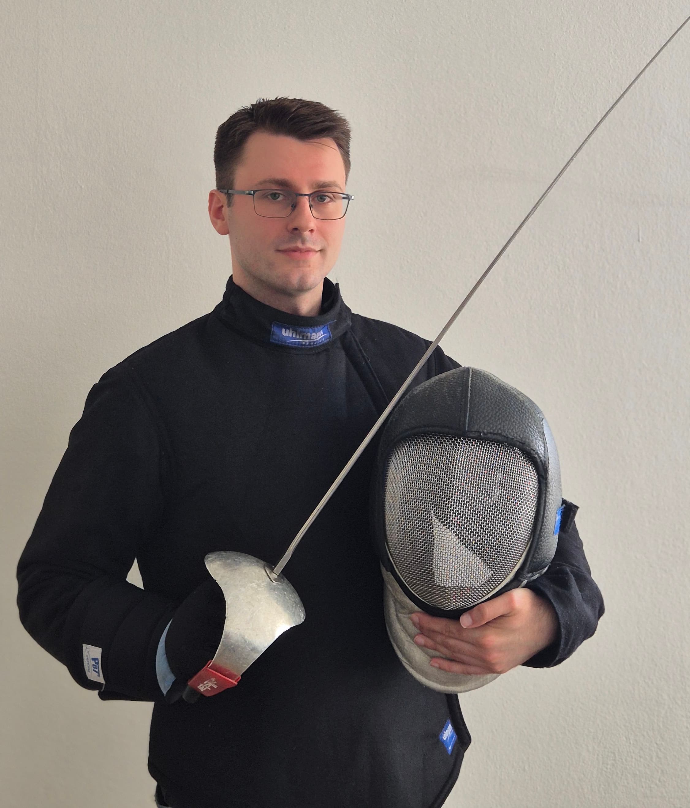 Photo von Nikola Kapralov, Trainer bei Riposte SU. Schwarze Kleidung, Säbel in der Hand, lächelnd.
